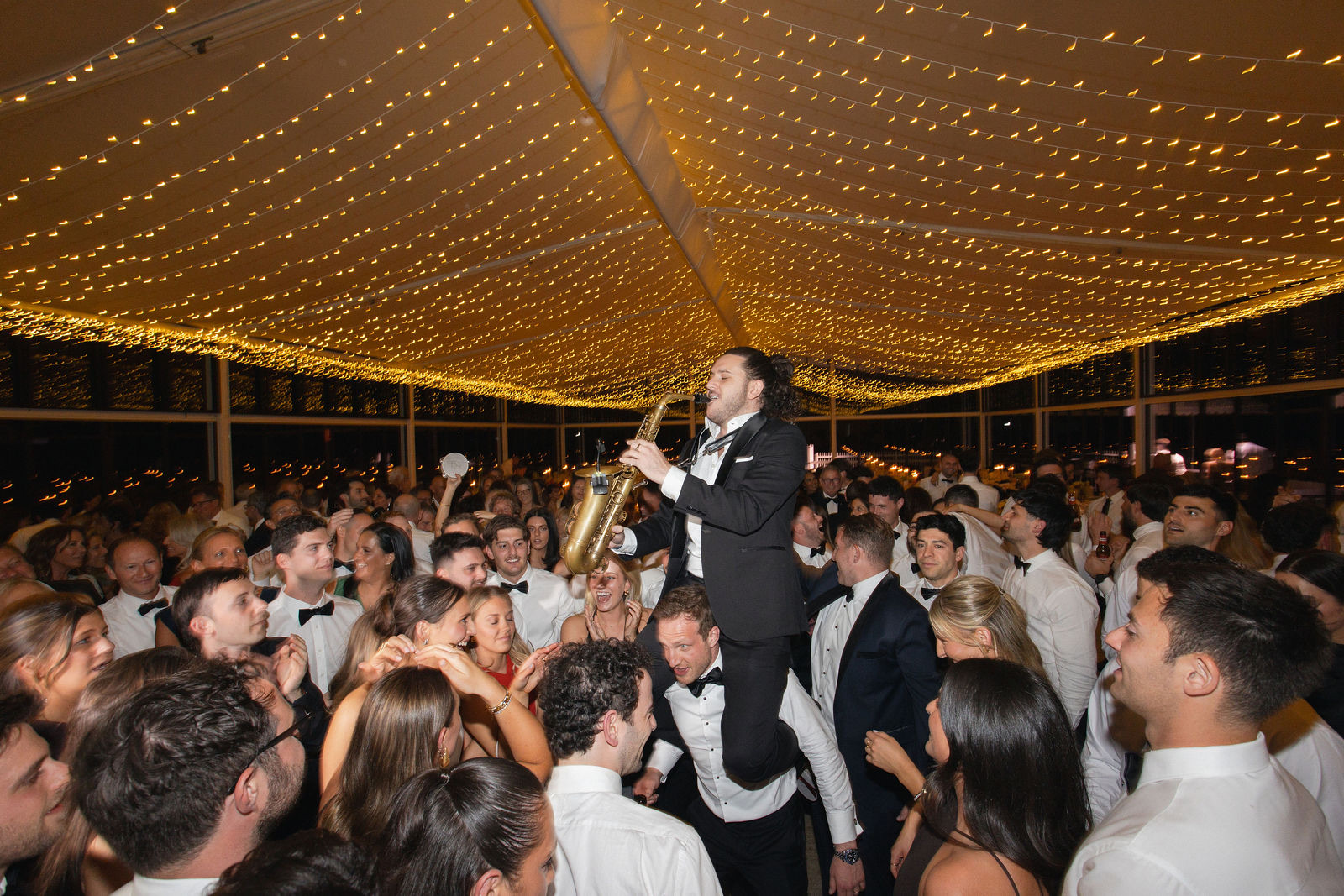 A saxophonist lifted above the dance floor under a marquee ceiling of fairy lights