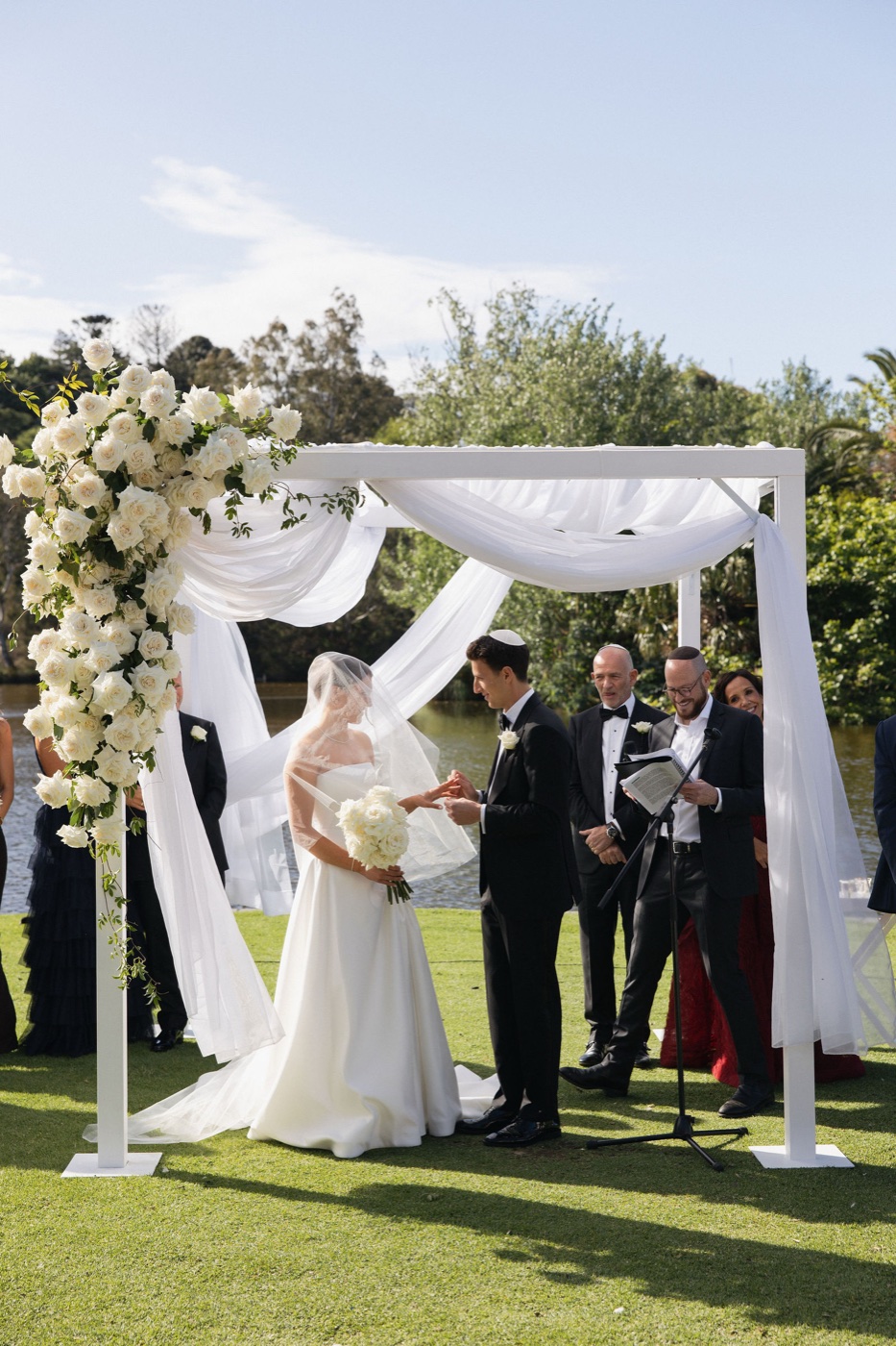 The couple exchanging rings under a white draped chuppah beside the lake