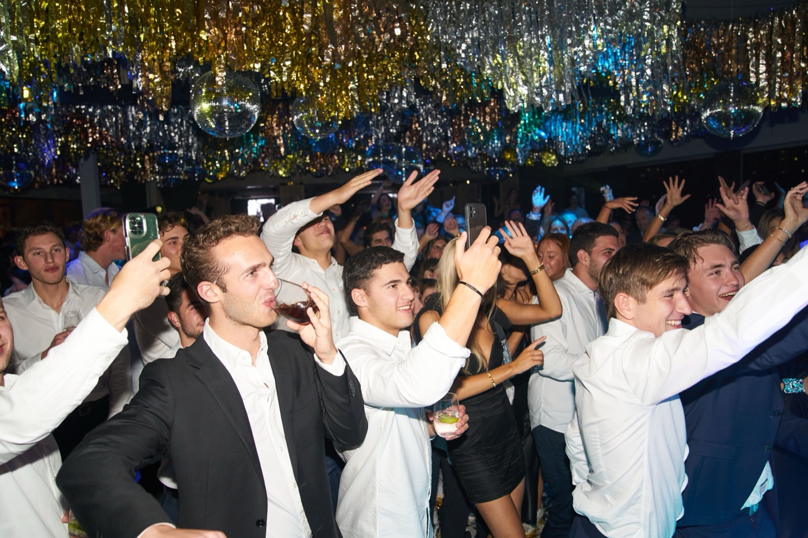 A packed party under disco balls and streamers, guests with their arms in the air