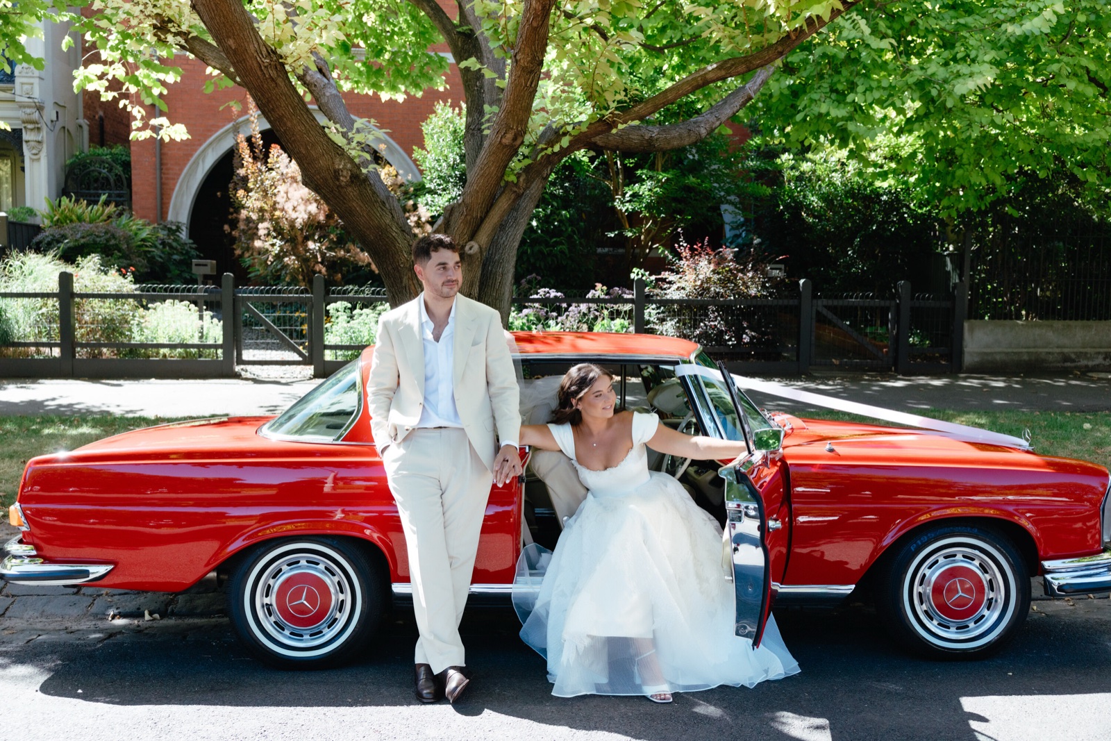 A bride and groom beside a vintage red car on a tree lined street